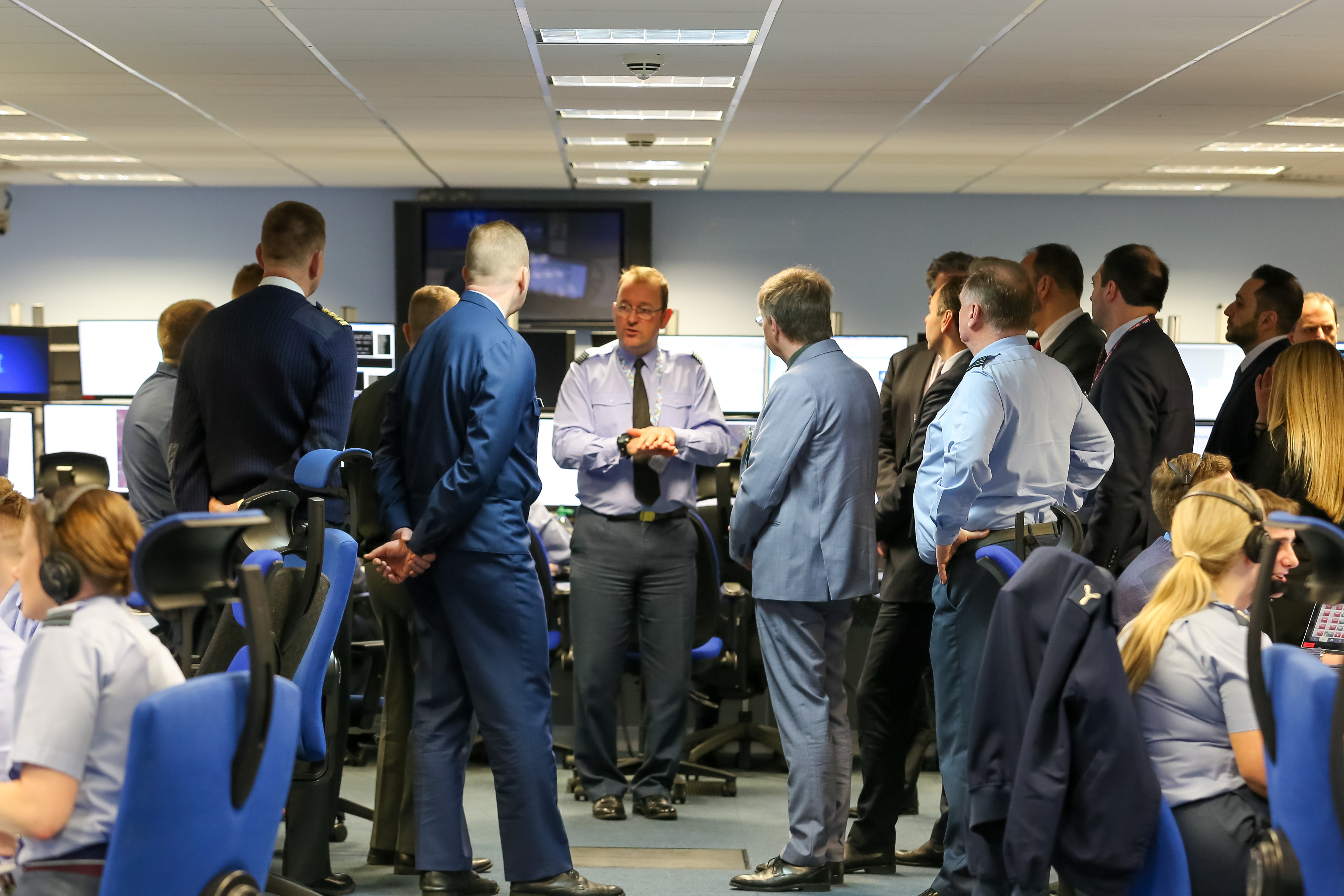 Image shows RAF aviators and civilians standing in discussion with some wearing headsets at desks.